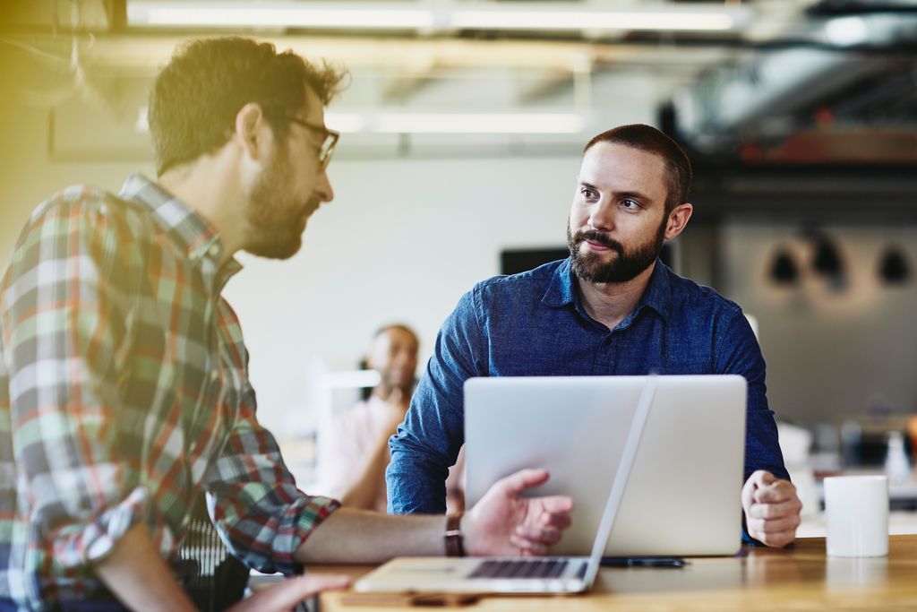 Two males talking. Both have beards. One wears glasses. One male in the background. Informal setting. Inside. Checkered shirt. Supporting color blue.