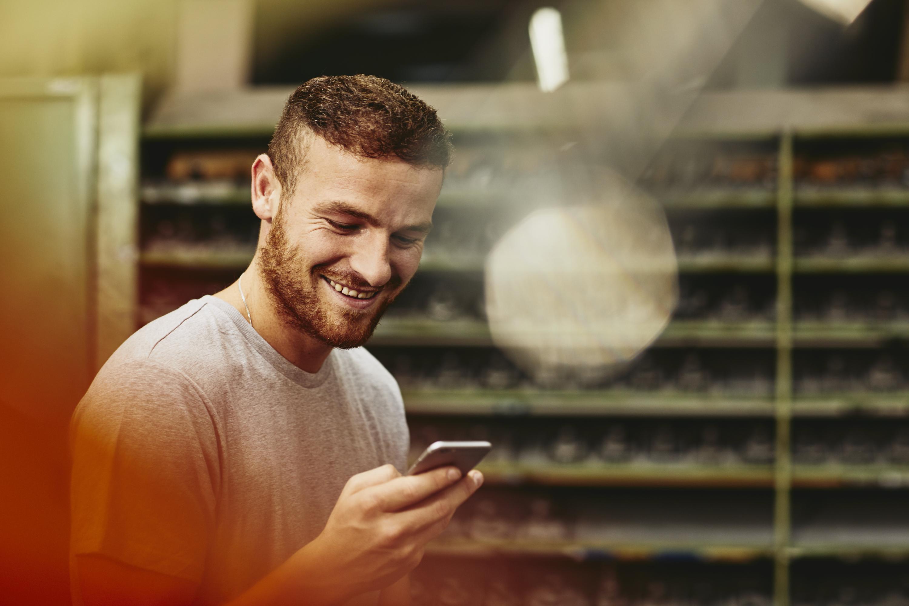 Blue-collar worker looking at his mobile phone. Tech environment. Smiling. Groomed beard. Greyish T-shirt. Primary color red.