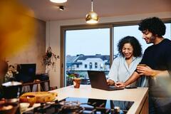 Woman and man standing next to the kitchen counter top, looking at a laptop. Smiling.