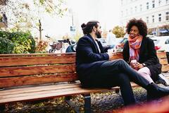Man and woman with drinks sitting on a bench outside while having a conversation.