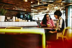 Two woman sitting in restaurant having a converstation, looking at laptop, smiling.