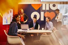 Group of business men and women in a meeting room with a phone, a screen and a laptop.