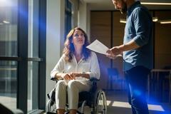 Kobieta na wózku inwalidzkim rozmawia z mężczyzną w biurze. Wygenerowano przez AI.