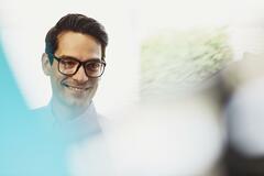 Male employee smiling. Caucasian man. Wearing glasses. Solid white shirt. Primary color cream. Secondary color blue.