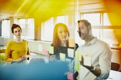Two business women and a man in an office putting sticky notes on a window. Primary colors: yellow and blue.