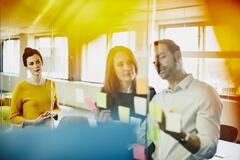 Two business women and a man in an office putting sticky notes on a window. Primary colors: yellow and blue.