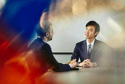 Two business men talking in a meeting, sitting, tablet, whiteboard in the background.