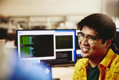 Man smiling while sitting behind his desk, computer screens displaying programming code.