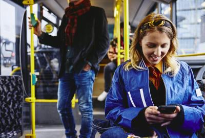 Smiling woman sitting in a bus, looking at her phone. Other people in the background.