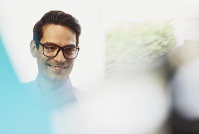 Male employee smiling. Caucasian man. Wearing glasses. Solid white shirt. Primary color cream. Secondary color blue.