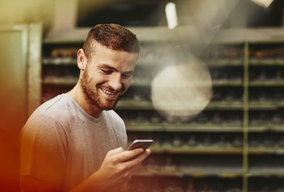 Blue-collar worker looking at his mobile phone. Tech environment. Smiling. Groomed beard. Greyish T-shirt. Primary color red.