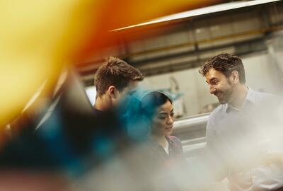 People in a tech environment discussing. Two males and one female. Caucasian (white) men and Asian woman. Primary color yellow. Secondary color blue.