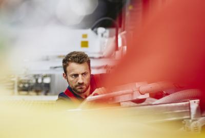 Blue-collar worker aligning something. Very focused. Caucasian man. Groomed beard. Primary color red. Secondary color cream/yellow.