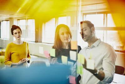 Two business women and a man in an office putting sticky notes on a window. Primary colors: yellow and blue.