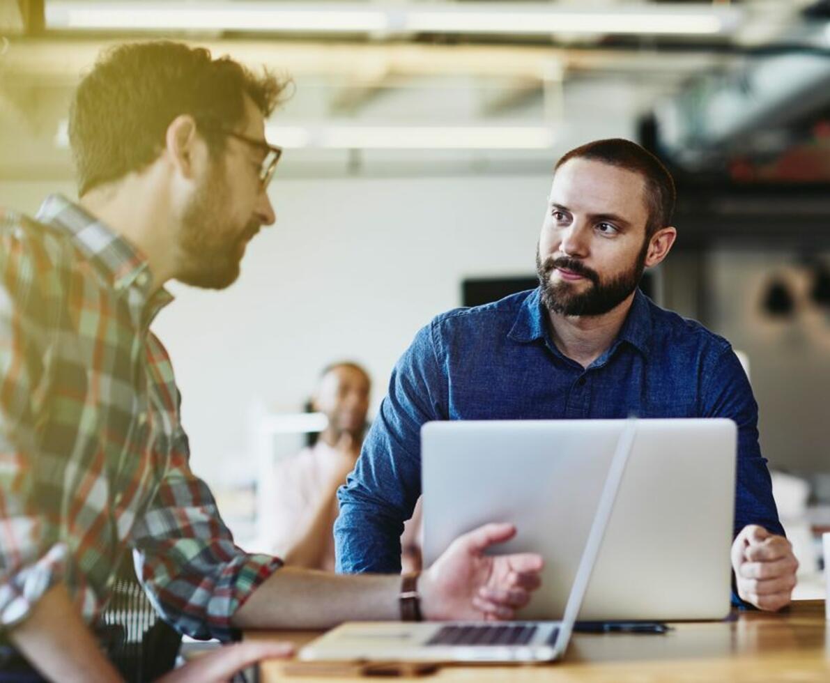 Two males talking. Both have beards. One wears glasses. One male in the background. Informal setting. Inside. Checkered shirt. Supporting color blue.