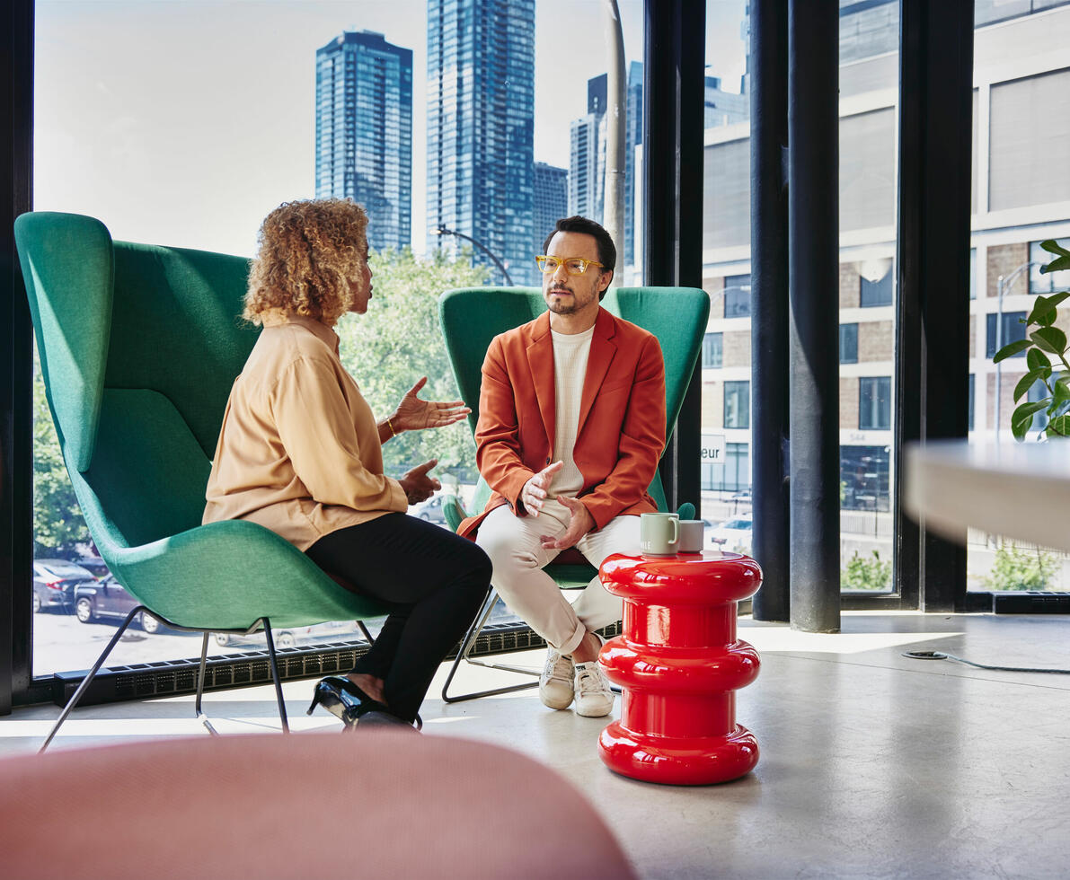 Male with glass and red blazer sitting in a green chair talking to a female sitting in a green chair