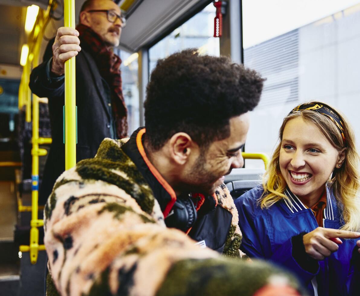 Woman and boy having a conversation, smiling, sitting in a bus. Looking at a phone. Other people in the background.