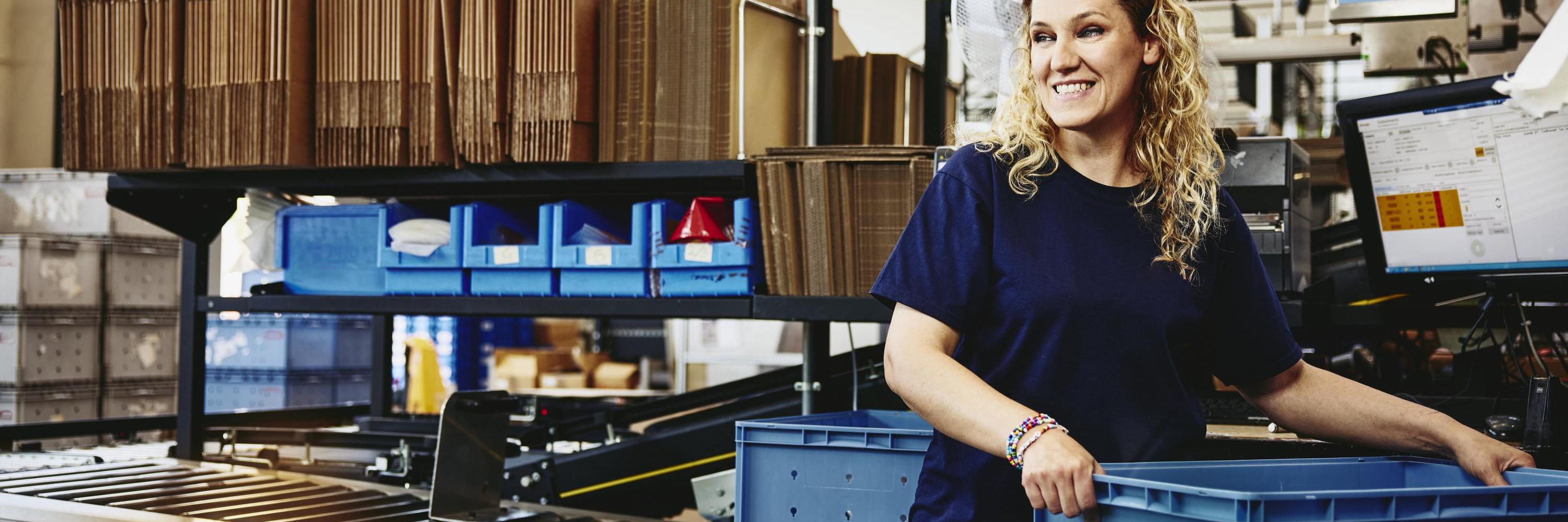 Smiling woman standing next to conveyer belt holding a crate