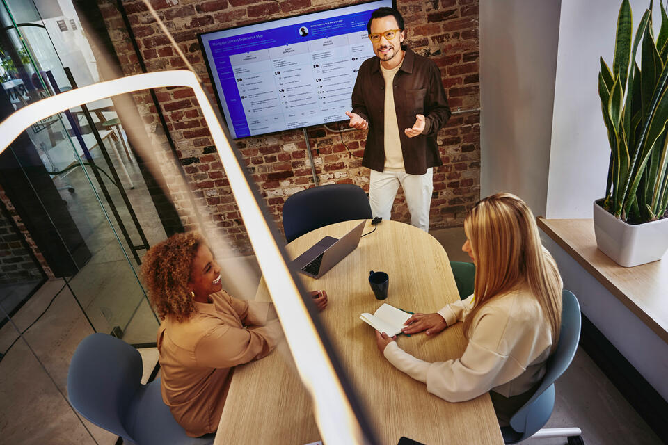 2 жінки сидять за столом і чоловік стоїть перед екраном і пояснює