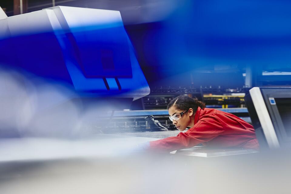 Працівниця-жінка щось вирівнює.  Афроамериканка.  Носіння захисних окулярів та комбінезону.  Зосереджений.  Кінський хвіст.  Загальна.  Основний колір синій.  Вторинний колір вид.