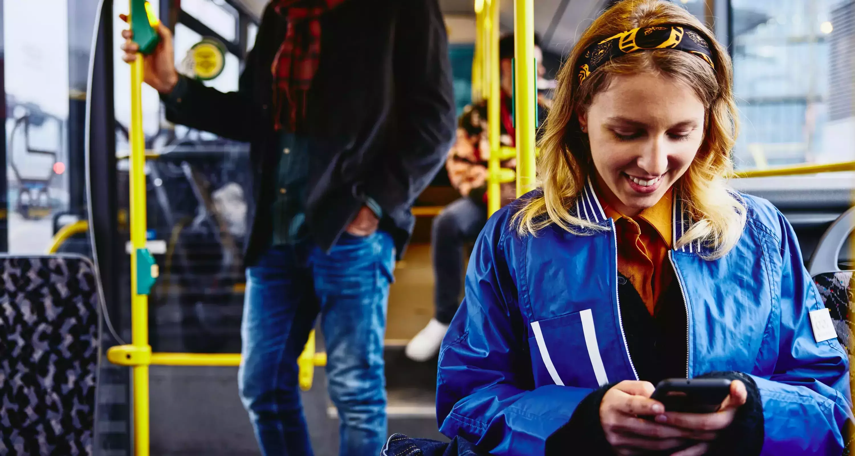 Smiling female sitting in a bus, looking at her phone. Other people in the background.
