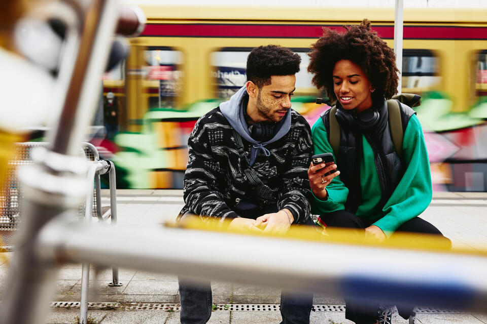 male and female looking at phone while sitting on a bench on a train platform.